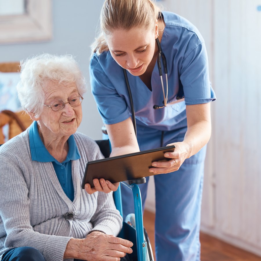Pflegefachkraft bespricht mit einer Seniorin im Rollstuhl Informationen am Tablet
